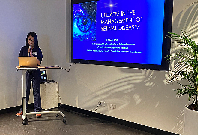 Dr Mei Tan speaking into a microphone standing beside a screen bearing the title slide of a PowerPoint presentation, entitled 'Updates in the management of retinal diseases.'