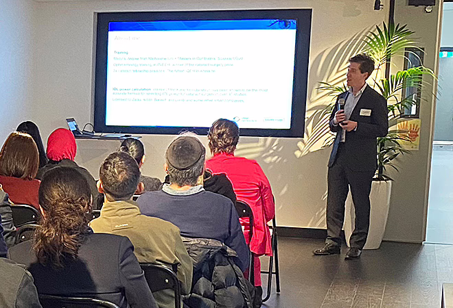 Dr Jack Kane speaking into a microphone while standing next to a screen onto which a Powerpoint presentation is projected, while audience members look on attentively.