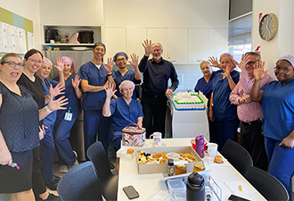 A dozen Windsor Gardens Day Surgery employees sitting and standing around a table in the break room, waving and grinning at the camera. Also visible is the Windsor Gardens Day Surgery cake and a selection of morning tea pastries.
