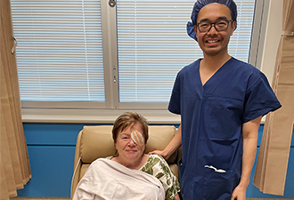 Dr Soo Khai Ng in scrubs standing next to Windsor Garden's 10,000th patient, Julie Spencer, who has just finished cataract surgery. She has a patch on her left eye. Both Dr Ng and Julie are smiling at the camera.