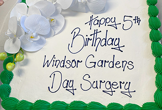 A close-up of the Windsor Gardens Day surgery birthday cake. The cake is rectangular and covered with white icing, with some decorative green piping around the edges and some artificial white orchids on the top left-hand side of the cake. 'Happy 5th Birthday Windsor Gardens Day Surgery' is written in purple icing on the cake.