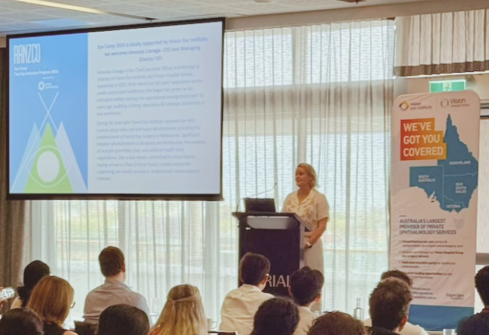 VEI CEO, Amanda Cranage's welcome address. She is standing in front of a crowd, positioned behind a lectern with a microphone. A projector screen is positioned to the left of the image. VEI signage is positioned to the right of the image.