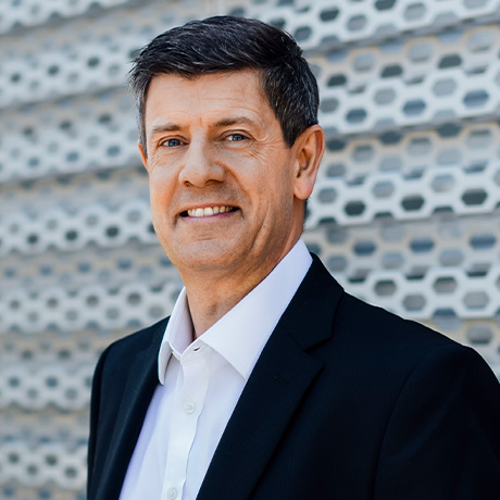 Headshot of A/Prof Tim Roberts in a black suit and white shirt (no tie) in front of a white textured feature wall outside Chatswood Day Surgery