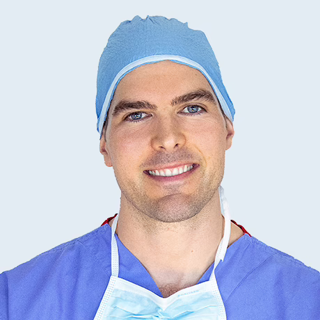 Headshot of ophthalmologist A/Prof Nick Andrew in blue scrubs, including scrub cap. There is an unused surgical face mask hanging around his neck.